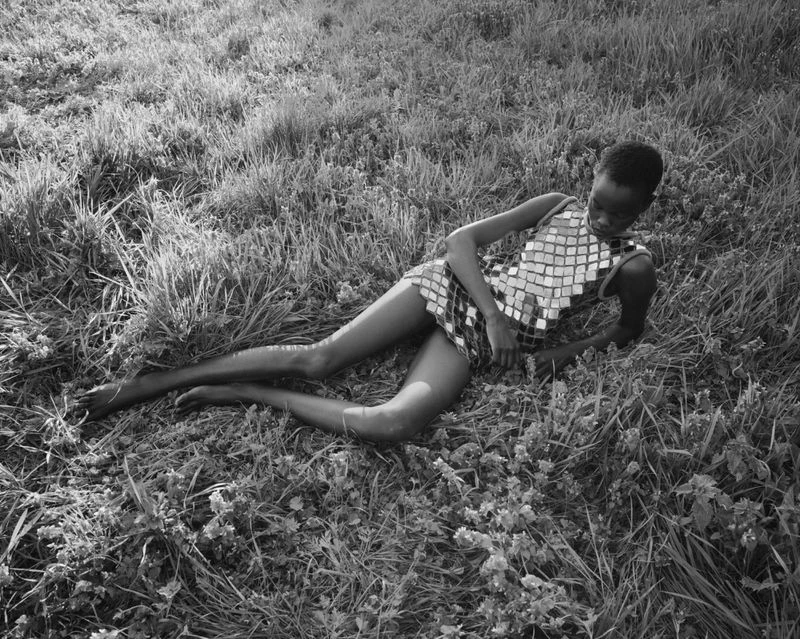 A black and white image shows a slender person in a mirrored mini dress reclining in tall grass in an open field under dappled sunlight.