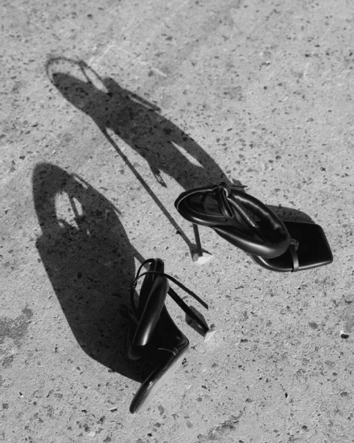 Two black heeled sandals sit on a textured concrete surface, casting a distinct shadow in a black-and-white fashion photograph.