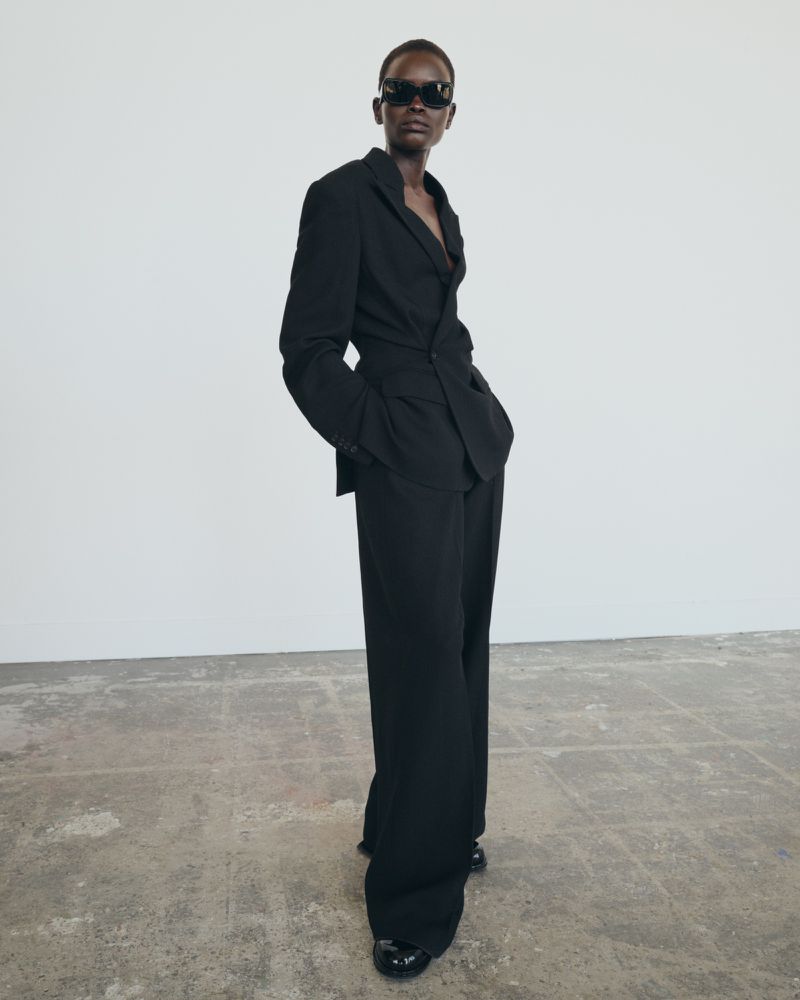 Full-length shot of a Black female model posing in a black suit with sunglasses, standing on a concrete floor against a white wall.