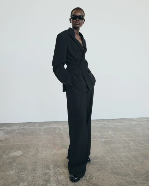 Full-length shot of a Black female model posing in a black suit with sunglasses, standing on a concrete floor against a white wall.