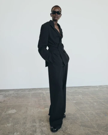 Full-length shot of a Black female model posing in a black suit with sunglasses, standing on a concrete floor against a white wall.