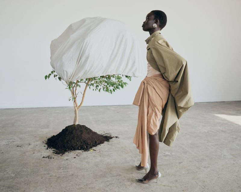 Fashion editorial shot of a Black woman in draped clothing posing next to a tree covered with a white cloth in a minimalist studio.