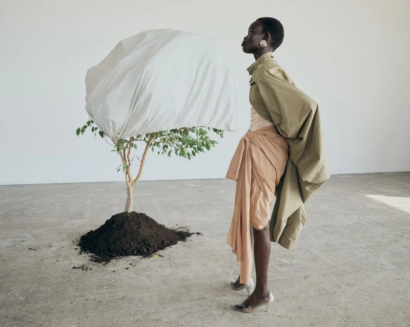 Fashion editorial shot of a Black woman in draped clothing posing next to a tree covered with a white cloth in a minimalist studio.