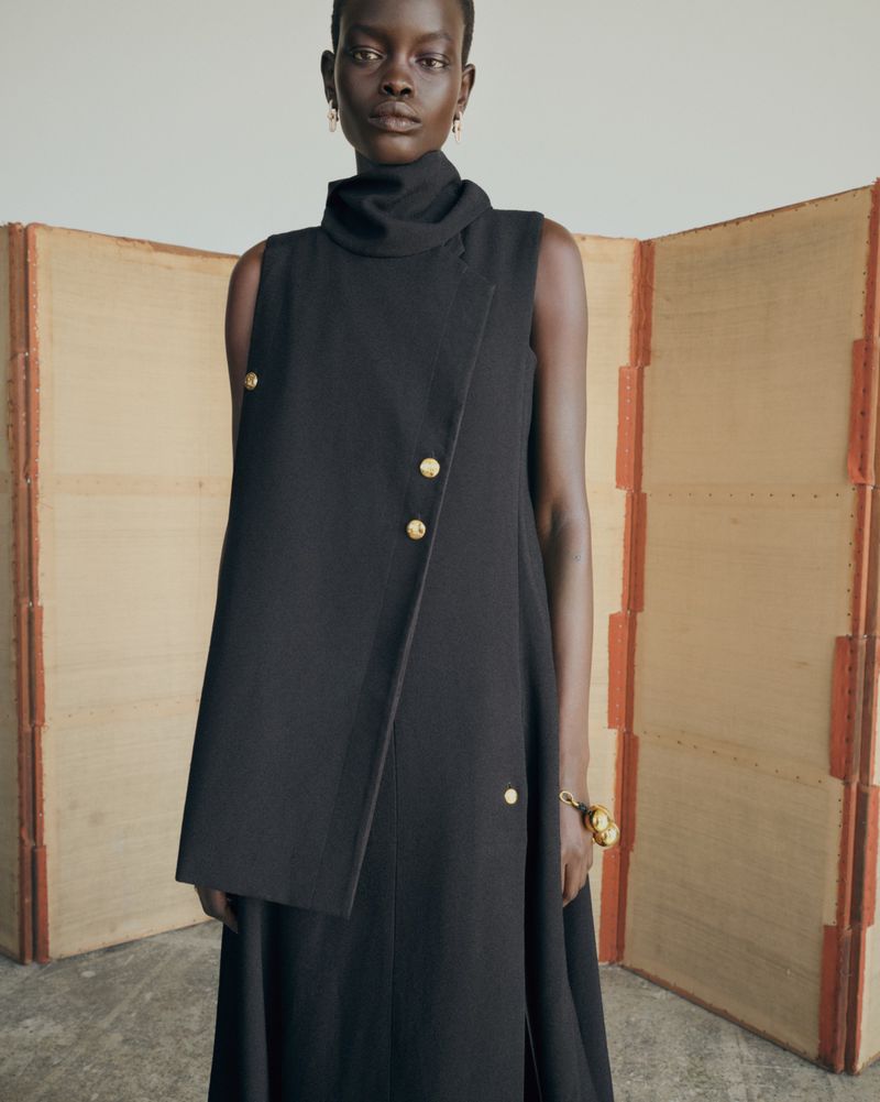 A woman in a black sleeveless dress stands against a neutral backdrop with a folding screen, presenting a minimalist fashion style.