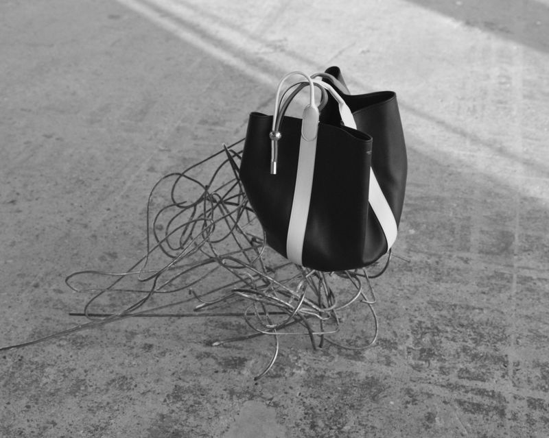 Black leather tote bag with white stripe on a concrete surface with silver wire decoration, creating a minimalist fashion still life.