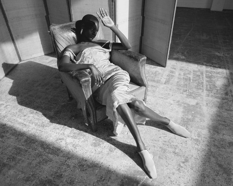 Elegant black-and-white image of an African woman seated in an armchair, wearing a light-colored dress and point shoes, with sunlit shadows.