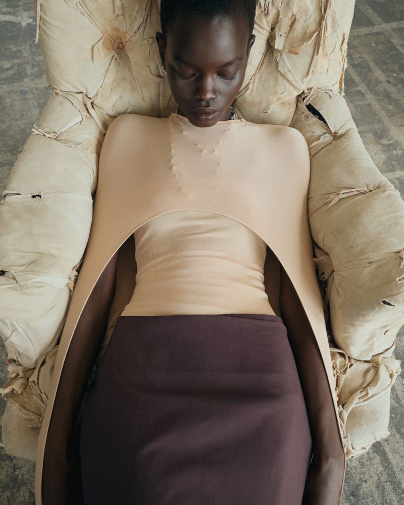 Model Athiec Geng reclining in a tattered cream armchair wearing a sculptural peach-beige top and dark brown skirt.