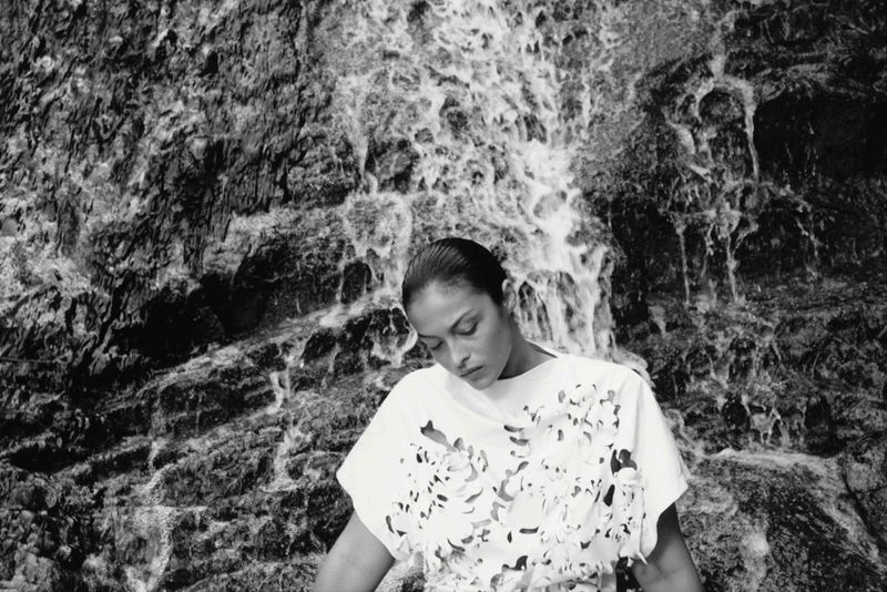 Monochrome portrait of an adult woman with her head tilted down in front of a rockface waterfall, wearing a white cut-out floral top.