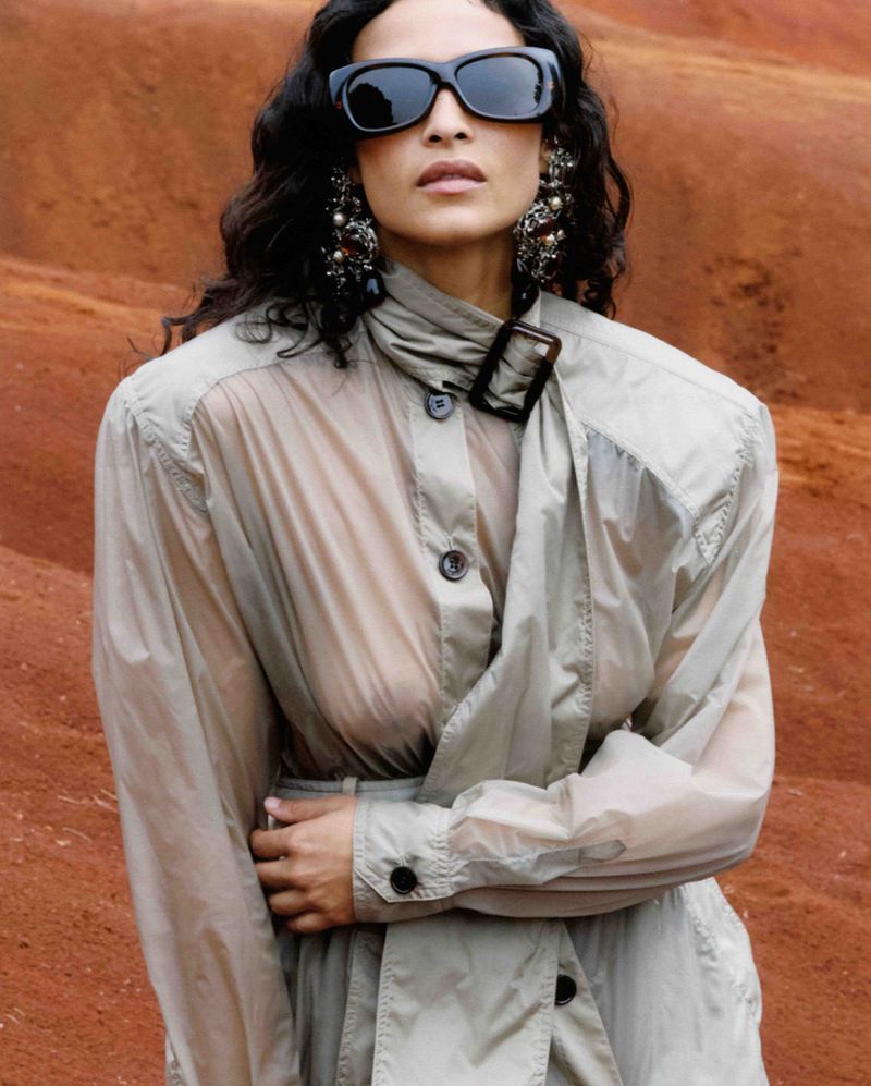 Woman wearing sunglasses and a tan trench coat poses against a reddish-brown desert backdrop, showcasing a fashion editorial style.
