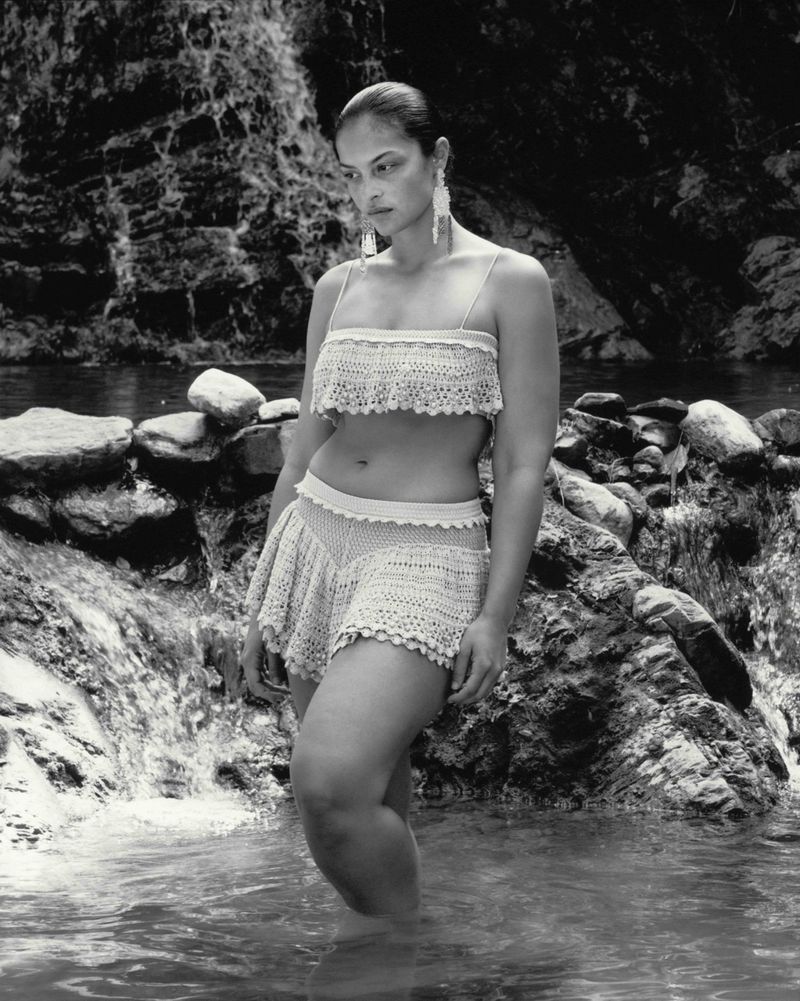 An adult female model poses standing in water, wearing a two-piece crochet set, with a tropical rock and waterfall background.