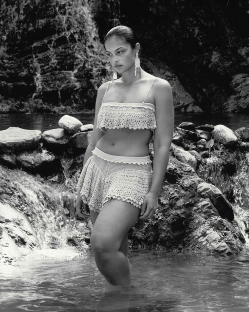 An adult female model poses standing in water, wearing a two-piece crochet set, with a tropical rock and waterfall background.