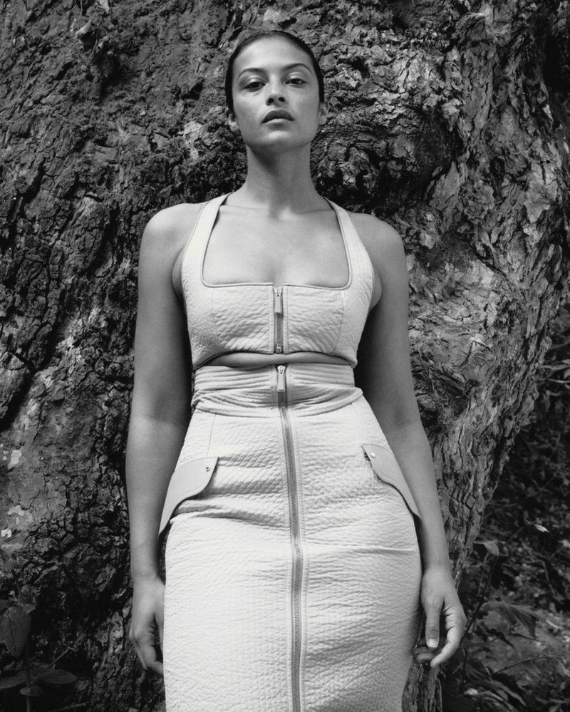 Monochrome portrait of a woman in a fitted top and skirt posing in front of a textured tree trunk in an outdoor setting.