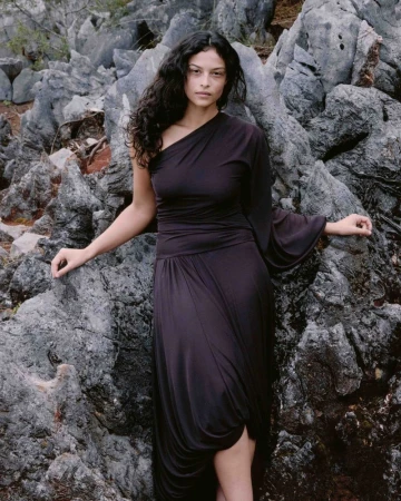 Model Devyn Garcia in a draped one-shoulder dark dress standing against a rugged grey rock formation.