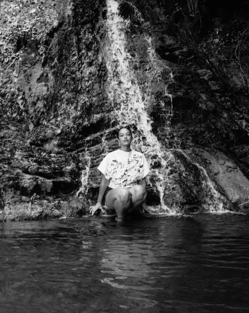 Monochrome portrait of a woman crouching in shallow water near a rocky waterfall, with a contemplative expression in a natural setting.