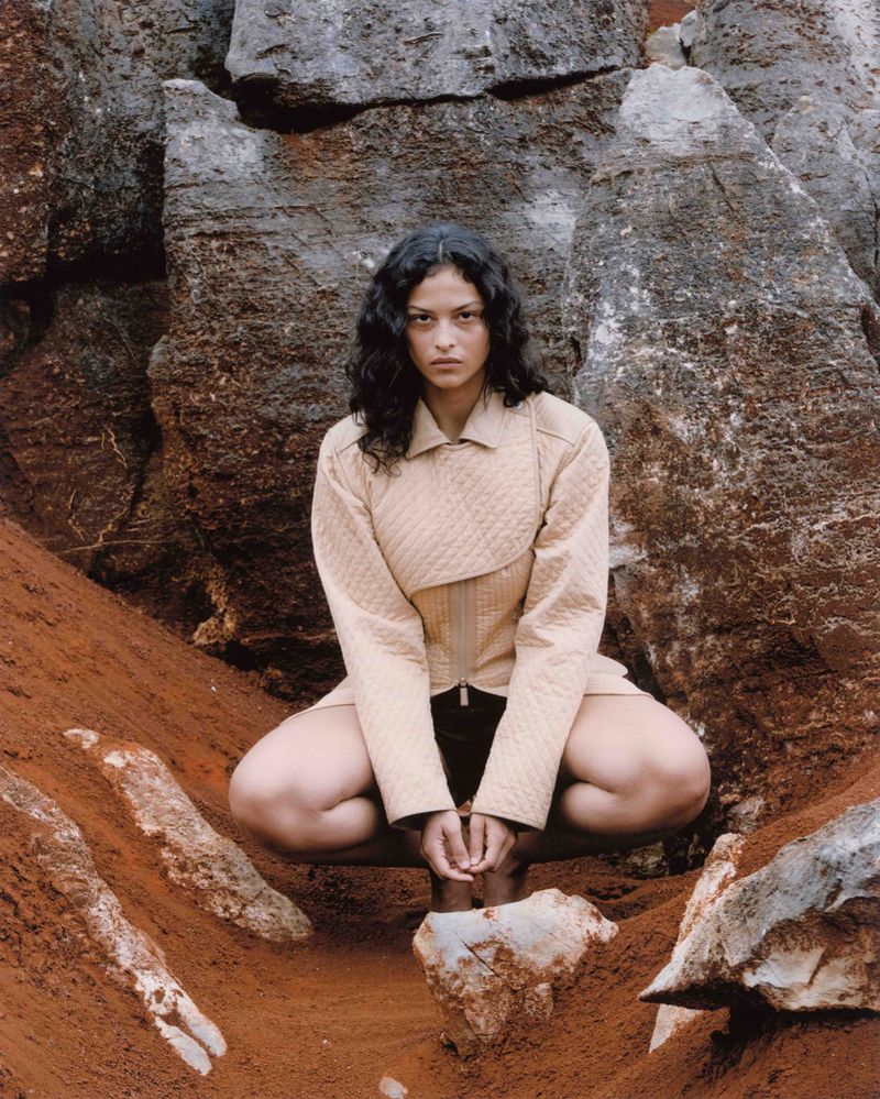 A woman in a beige jacket and skirt poses in a crouching position among red dirt and large rock formations in an outdoor setting.