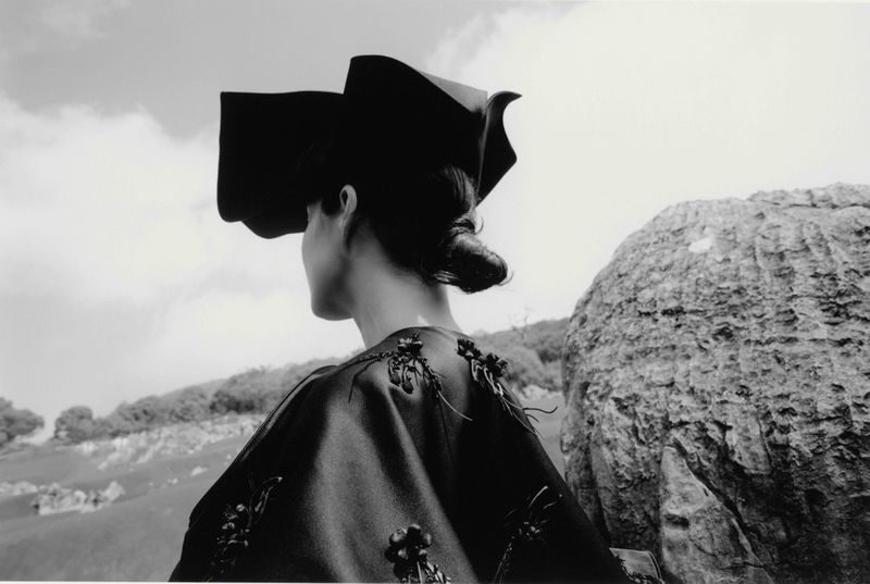 Black-and-white outdoor fashion shot: Back view of an adult woman wearing a unique dark hat and decorative top, with a rocky landscape.