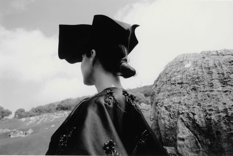 Black-and-white outdoor fashion shot: Back view of an adult woman wearing a unique dark hat and decorative top, with a rocky landscape.