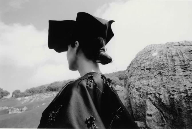 Black-and-white outdoor fashion shot: Back view of an adult woman wearing a unique dark hat and decorative top, with a rocky landscape.