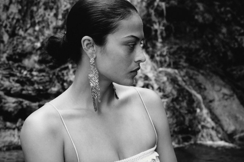 Woman with dark hair in a bun wearing dangling statement earrings in a monochrome portrait, posing in front of a nature scene.