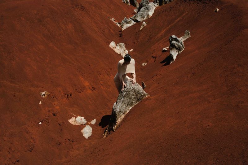 Adult female climbs a rock formation surrounded by a red desert landscape. The sunlight illuminates the woman and the terrain.