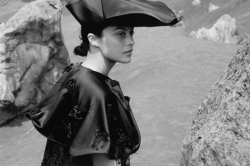 Monochrome fashion shot of a woman in a dramatic hat and beaded cape, posing amongst rocks in a high-contrast outdoor setting.