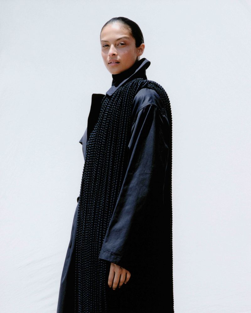 Model Devyn Garcia posing in a long black overcoat and a textured draped black fringe accessory against a plain white background.