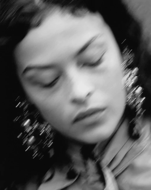 Close-up portrait of an adult woman with dark curly hair wearing statement earrings. The image has a soft focus and monochromatic colour grade.