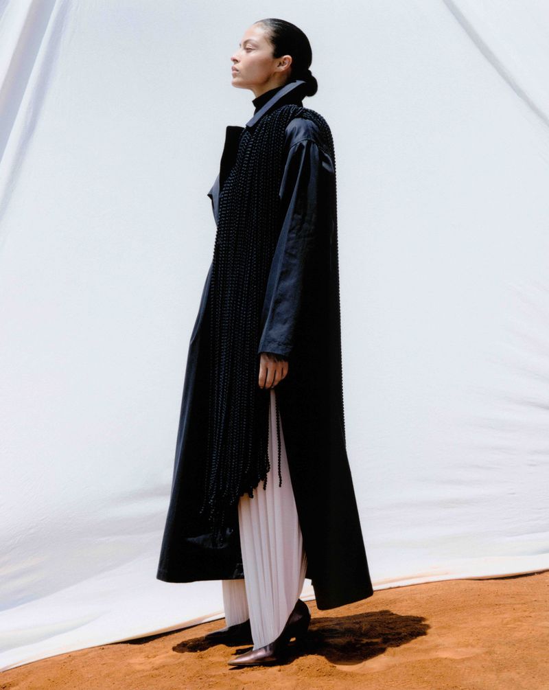 A woman in a long black coat and white pleated pants stands against a white backdrop with reddish-brown ground. Fashion shot.