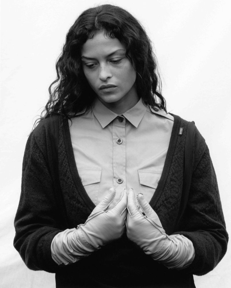 Monochrome portrait of a woman with long curly hair wearing fingerless gloves, a button-down shirt, and a dark cardigan, looking down.