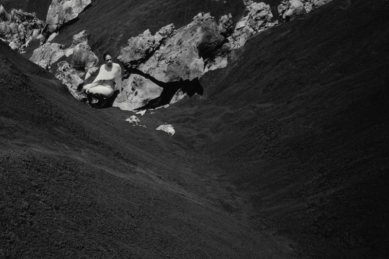 Adult female in white clothes seated in meditation pose on slope with dark moss and rocks, outdoor, black and white.