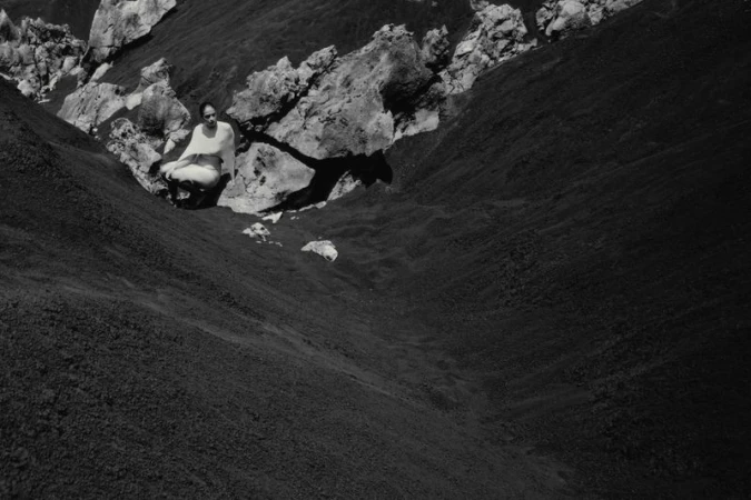 Adult female in white clothes seated in meditation pose on slope with dark moss and rocks, outdoor, black and white.