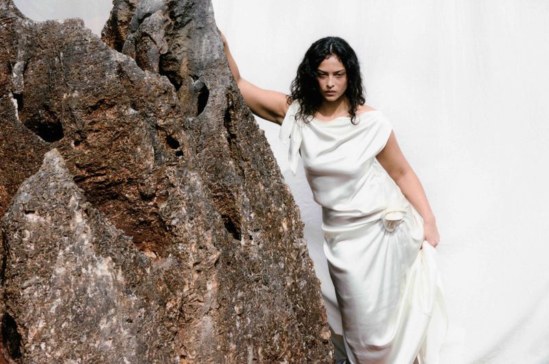 Woman in a white gown poses next to a large rock formation against a white background, portrait framing, studio lighting. 