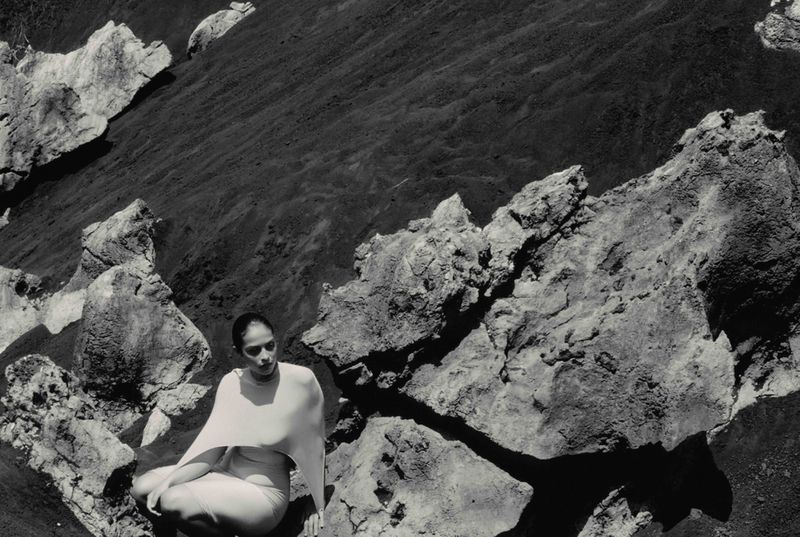 Black and white photograph of an adult woman wearing a white dress, seated among large gray rocks. Dark volcanic sand in background.