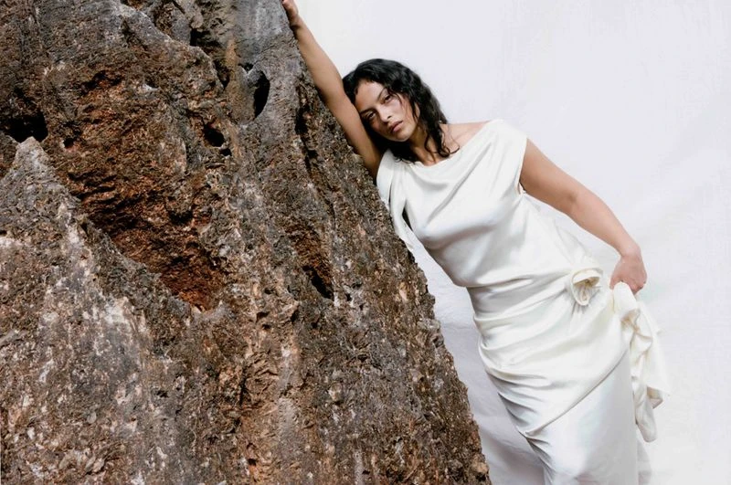Woman in elegant white dress leans against a textured rock formation with a serene expression against a bright, minimalist background.
