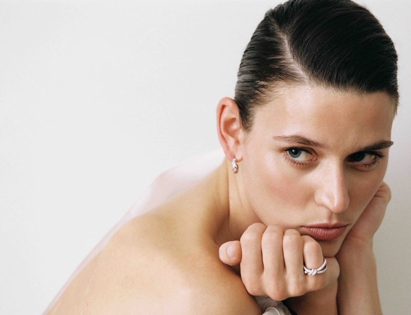 Close-up portrait of Vivienne Rohner with slicked-back dark hair, resting her chin on her hand, wearing delicate silver jewelry.