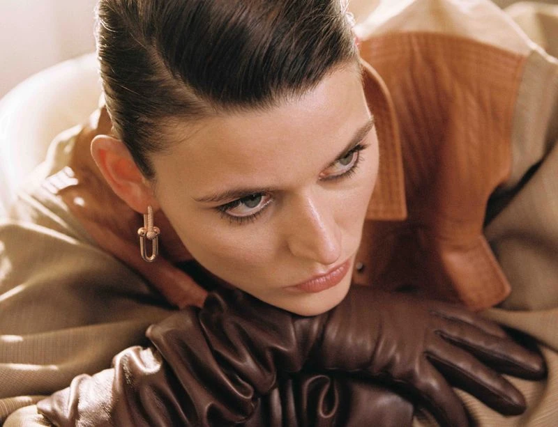 Eye-level close-up of a poised adult woman with slicked-back hair, showcasing fashion earrings and gloves, set against a neutral backdrop.