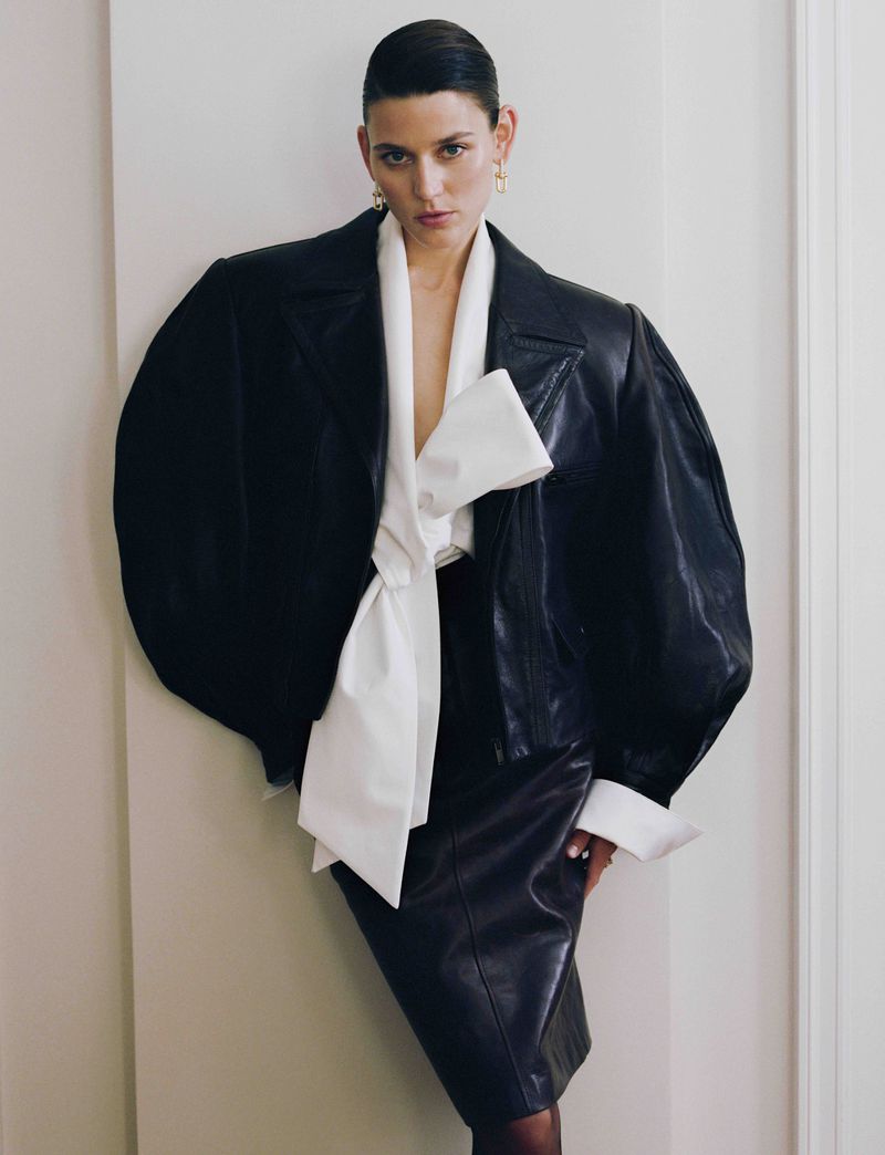 Model Vivienne Rohner in a black oversized leather jacket with balloon sleeves and a large white bow top against a white wall.