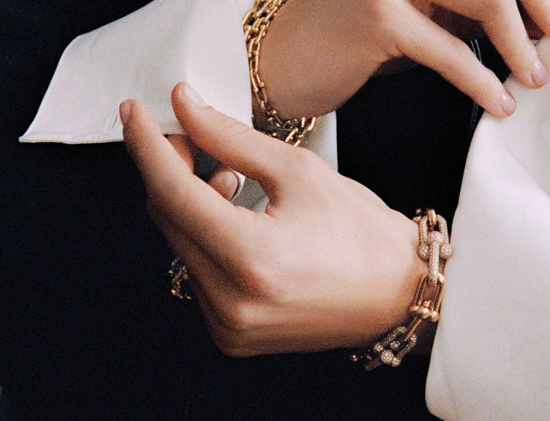 Close-up of a woman's hands, adorned with gold link bracelets, set against a backdrop of a white collared shirt and a black jacket.