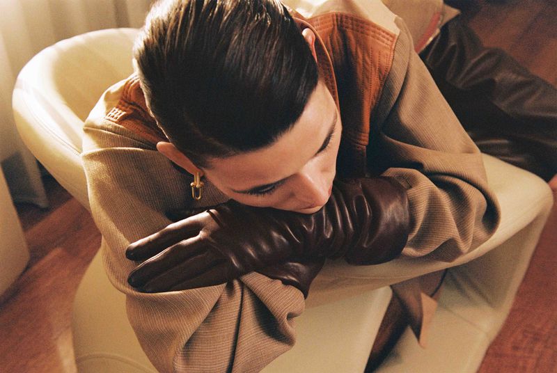 Adult with groomed dark hair leans on a cream armchair, wearing leather gloves and a tan jacket with an earring, indoor setting.