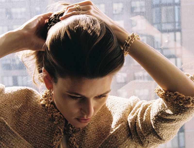 Vivienne Rohner adjusting her hair, wearing a textured tan jacket and gold jewelry, viewed against an urban window backdrop.