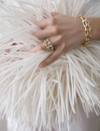 A model's hand wearing a gold link ring and bracelet rests atop a voluminous white feathered or fringed garment.