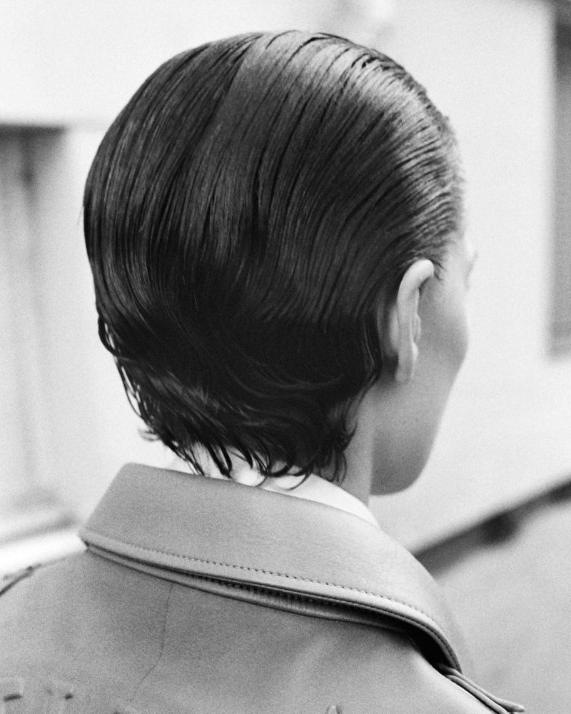 Rear view of a model with sleek dark wet hair wearing a collared coat in a black and white editorial portrait photograph