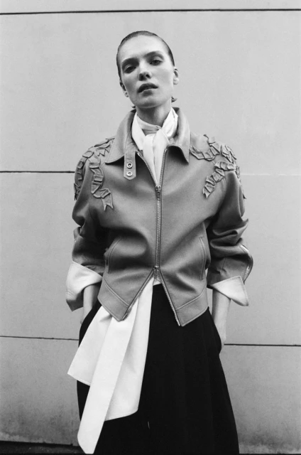 A young adult female model poses in a leather jacket with unique appliqués, paired with wide-legged trousers, set against a neutral wall backdrop.