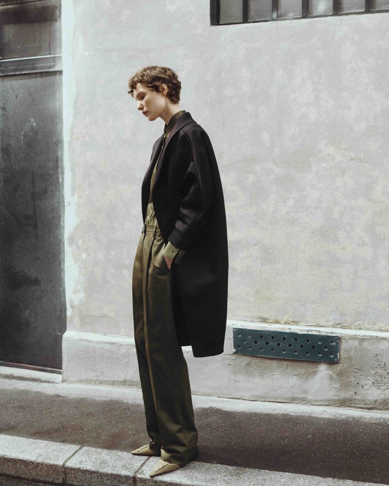 Fashion shot of a curly-haired model in a black coat, green shirt and pants, and gold shoes, posing outdoors against an off-white wall.