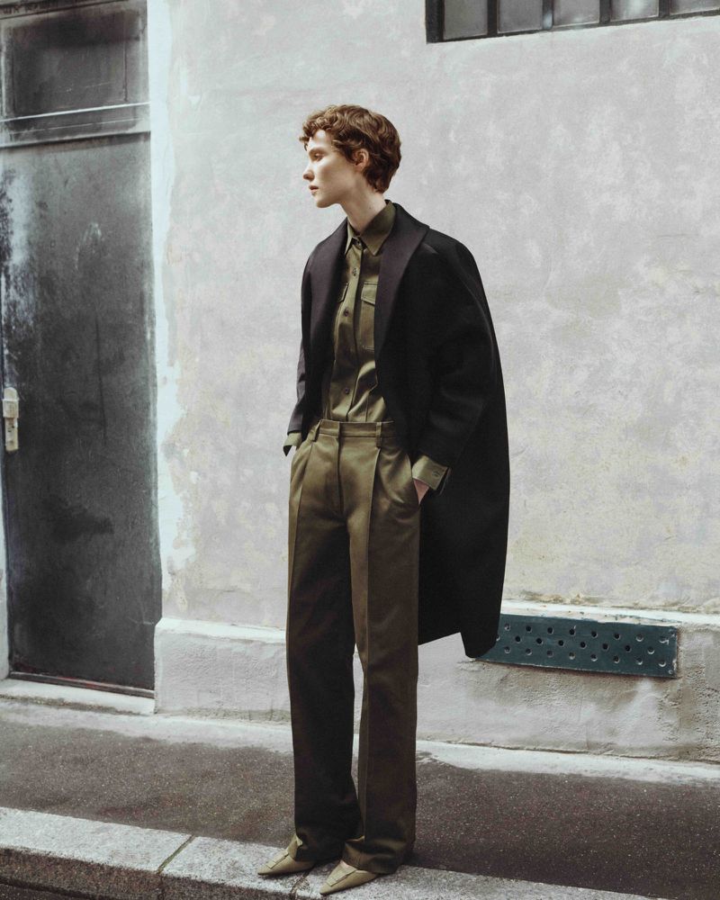 Fashion photo of a slim adult with reddish-brown hair in an olive-colored shirt and pants with a black coat, standing outdoors.