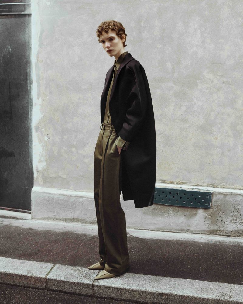 Adult woman poses in long black coat, olive shirt and pants, and tan shoes on a city sidewalk by a textured concrete wall.