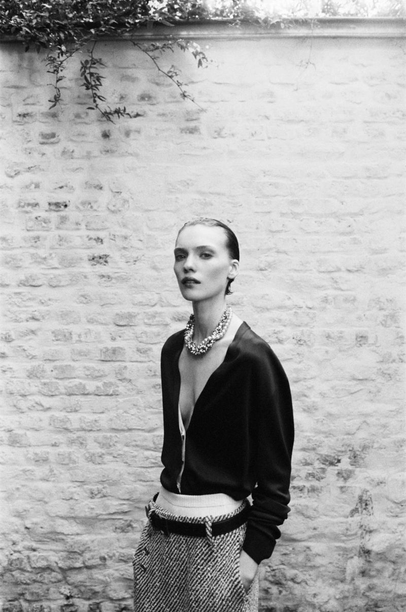 Black and white portrait of a female fashion model in a cardigan and skirt with a brick wall and foliage in the background.