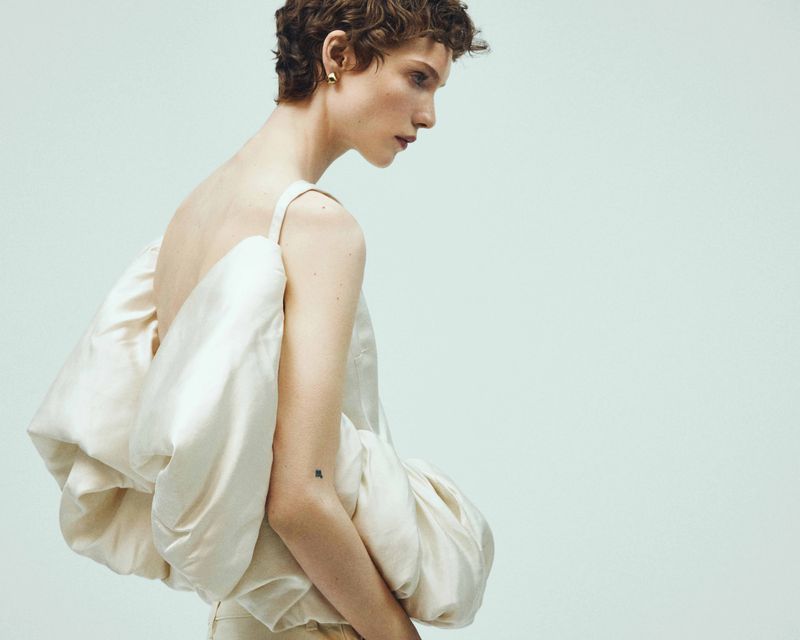 Adult model in a side-view portrait wears a strapless top with exaggerated folds and light-colored pants against a light background.