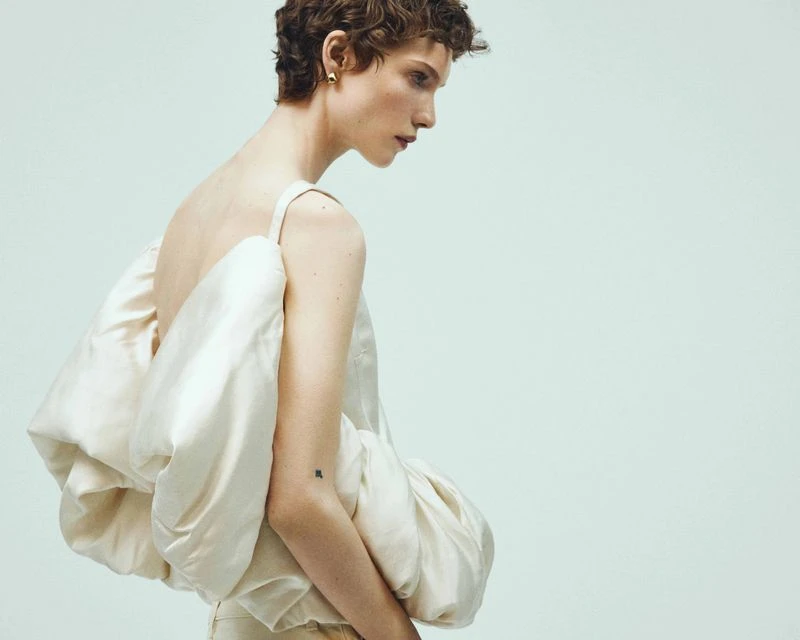 Adult model in a side-view portrait wears a strapless top with exaggerated folds and light-colored pants against a light background.