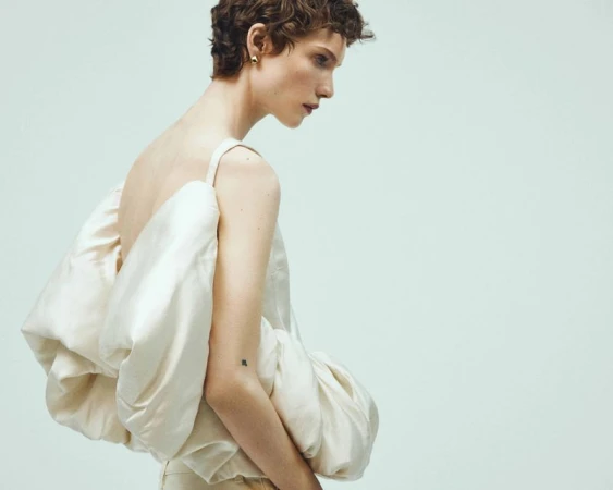 Adult model in a side-view portrait wears a strapless top with exaggerated folds and light-colored pants against a light background.